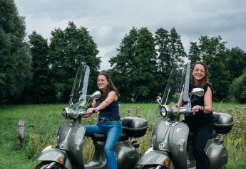 Deux femmes souriantes sur des scooters devant un paysage vert à la tiny house The Nutshell, Pays-Bas.