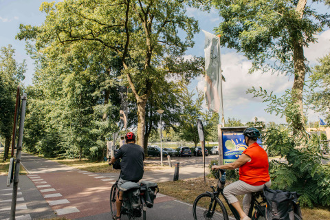 Two cyclists ride near the entrance to Recreation Park Den Blanken in the Netherlands, surrounded by trees.