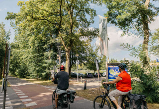 Two cyclists ride near the entrance to Recreation Park Den Blanken in the Netherlands, surrounded by trees.