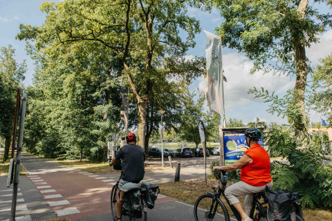 Twee fietsers aan de entree van recreatiepark Den Blanken in Nederland, geflankeerd door bomen.