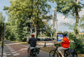 Twee fietsers aan de entree van recreatiepark Den Blanken in Nederland, geflankeerd door bomen.