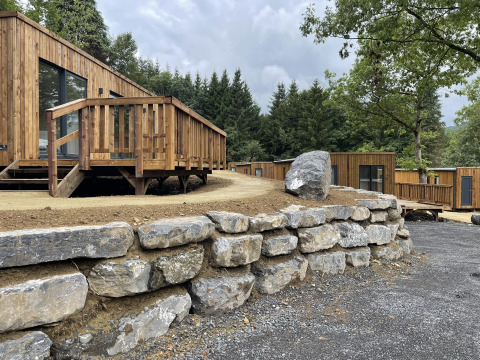 Lodges en bois modernes avec un muret en pierre, entourés d'arbres verdoyants dans un cadre forestier paisible.