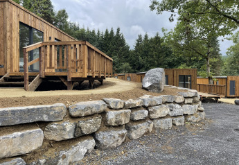Holzhütten mit Veranda und steiniger Mauer, umgeben von Wald und Bäumen, unter bewölktem Himmel.
