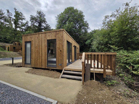 Een modern houten lodge met terras, midden in de natuur, omringd door bomen en een grindpad.