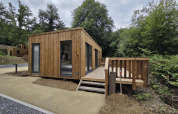 Eine moderne Holzhütte mit Terrasse mitten in der Natur, umgeben von Bäumen und einem Kiesweg.