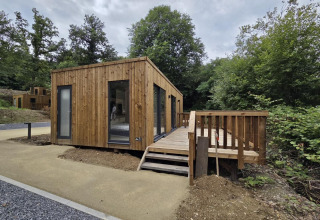 A modern wooden lodge with a terrace set in nature, surrounded by trees and a gravel path in front.