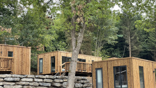 Modern wooden lodge cabins set on a wooded hillside, surrounded by lush green trees and stone walls.