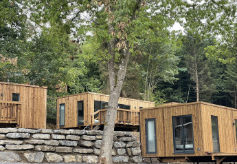 Moderne houten lodges aan een beboste heuvel, omringd door groene bomen en natuurstenen muren.