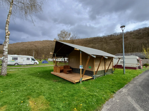 Tienda safari en Camping la Roche, Bélgica, con terraza de madera y rodeada de autocaravanas y vegetación.