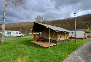 Tienda safari en Camping la Roche, Bélgica, con terraza de madera y rodeada de autocaravanas y vegetación.