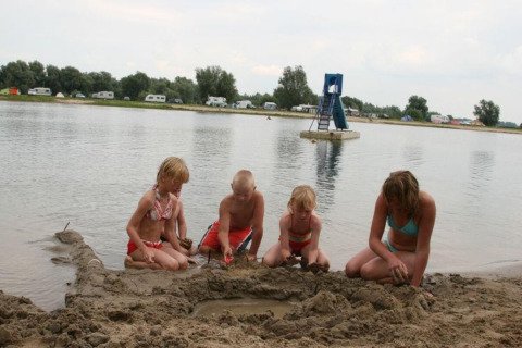 Kinderen bouwen een zandkasteel aan het water bij Recreatiepark de Scherpenhof met caravans op de achtergrond.
