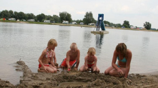 Børn leger i sandet ved søbredden ved Recreatiepark de Scherpenhof med campingvogne i baggrunden.