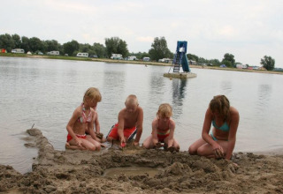 Kinderen bouwen een zandkasteel aan het water bij Recreatiepark de Scherpenhof met caravans op de achtergrond.