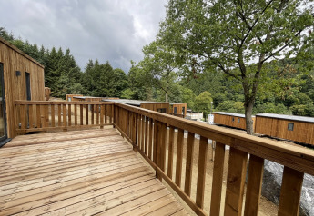 Houten terras van een lodge met uitzicht op houten huisjes, bomen en een bosrijke heuvelachtige omgeving.