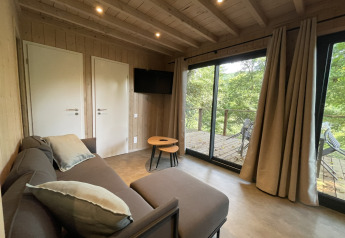 Soggiorno accogliente in un lodge con divano, tavolino, TV e grandi vetrate su terrazzo tra gli alberi.