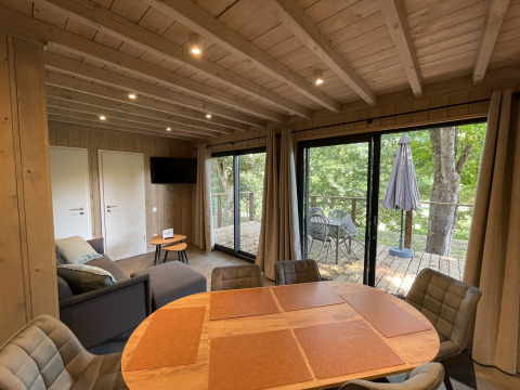 Intérieur de lodge lumineux avec table à manger, canapé, TV et accès à la terrasse surplombant la forêt.