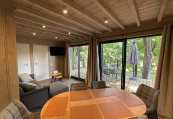 Lyst lodgeinteriør med spisebord, sofa og tv, der åbner ud til en terrasse med havemøbler og udsigt til skov.