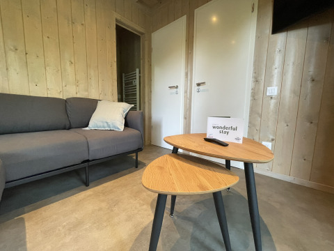 Gemütlicher Aufenthaltsraum in einer Lodge mit grauem Sofa, zwei Holztischen und Willkommensschild.