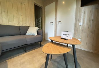 Gemütlicher Aufenthaltsraum in einer Lodge mit grauem Sofa, zwei Holztischen und Willkommensschild.