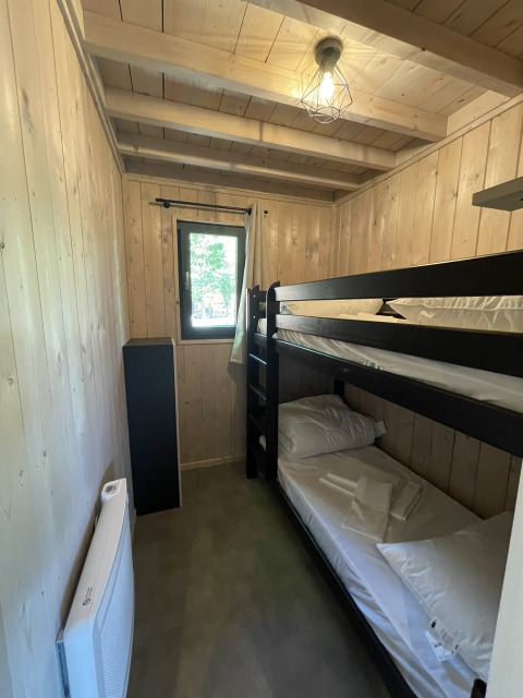 A small lodge bedroom with wooden walls, a black bunk bed, window, compact heater, and linens.