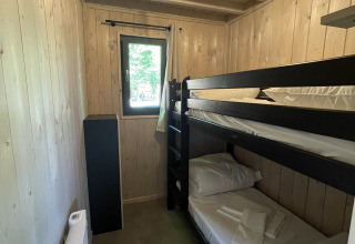A small lodge bedroom with wooden walls, a black bunk bed, window, compact heater, and linens.