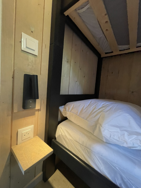 Gros plan sur un lit superposé à côté d’un mur avec prise, petite étagère et lampe de lecture dans un lodge.