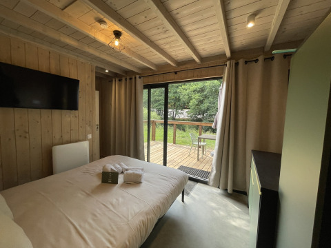 Knusse lodge slaapkamer met houten wanden, tweepersoonsbed, tv en zicht op terras en groene tuin.