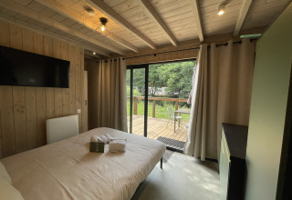 Knusse lodge slaapkamer met houten wanden, tweepersoonsbed, tv en zicht op terras en groene tuin.