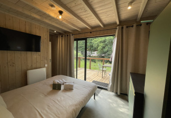 Knusse lodge slaapkamer met houten wanden, tweepersoonsbed, tv en zicht op terras en groene tuin.