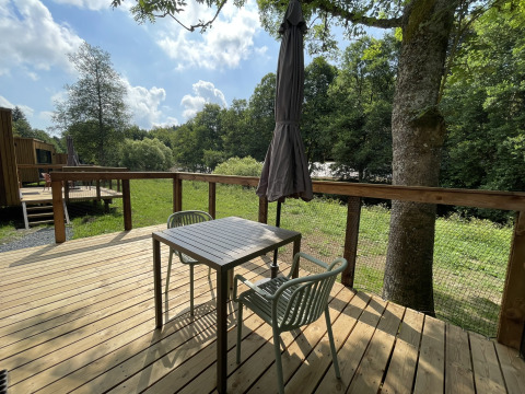 Zonnig houten terras aan een lodge met tafel, twee stoelen, parasol en zicht op gras en bomen.