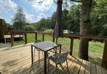 Sunny wooden deck at a lodge with a table, two chairs, an umbrella, and a view of grass and trees.