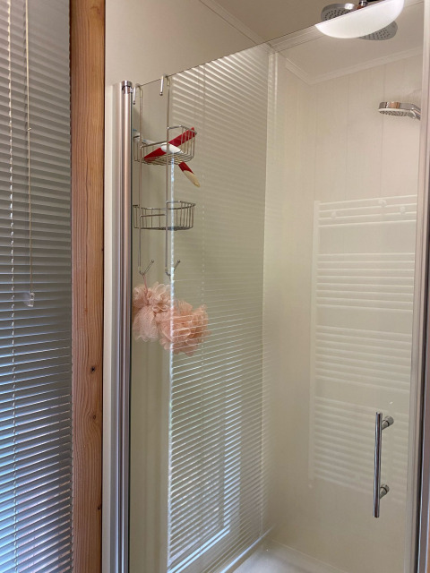 Cabina doccia con porta in vetro, cestino portaoggetti e spugna rosa, persiane riflesse sullo sfondo.