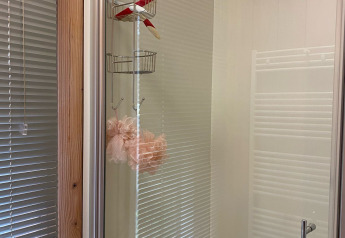 Duschkabine mit Glastür, Aufbewahrungskorb und rosa Duschschwamm, reflektierende Jalousien im Hintergrund.