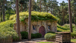 Hobbit huisje i skoven med stengulv og græstørvtag, omgivet af grønne træer og buske.
