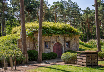 Cabaña Hobbit huisje de piedra con techo verde, en medio del bosque y vegetación exuberante.