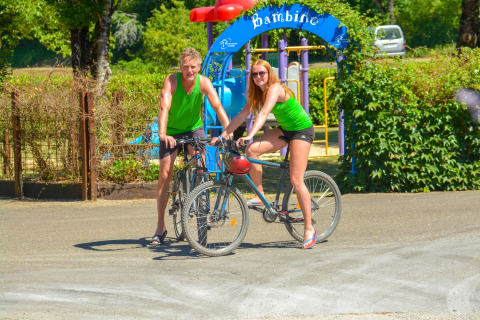 Twee mensen in groene sportkleding fietsen bij een speelplein in het vakantiepark La Draille, Occitanië, Frankrijk.