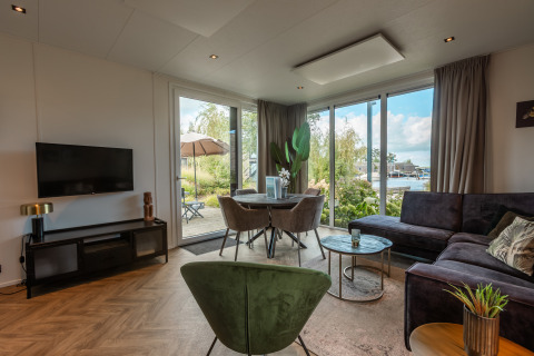 Salon moderne avec grandes fenêtres, canapé, fauteuil vert et vue sur l’eau au Vakantiechalet comfort+.