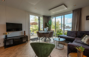Modernes Wohnzimmer mit großen Fenstern, Sofa, grünem Stuhl und Blick auf Wasser im Vakantiechalet comfort+.