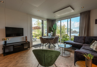 Soggiorno moderno con ampie finestre, divano, poltrona verde e vista sull’acqua al Vakantiechalet comfort+.