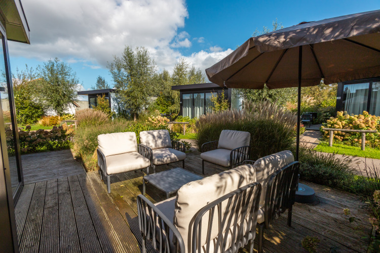 Terrasse einer Lodge mit gemütlichen Stühlen, Sonnenschirm und viel Grün in Vakantiepark De Wiedense Meren.