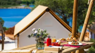 Petit-déjeuner sur une table en bois devant une tente safari avec vue sur l’eau et la verdure autour.