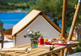 Colazione su un tavolo di legno davanti a una tenda safari con vista su acqua e natura verdeggiante.