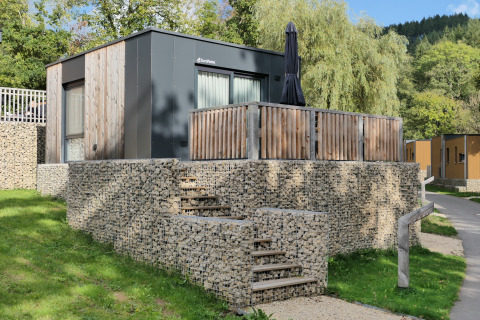 Moderne tiny house met houten gevel, stenen trap en terras, omringd door groen en weelderige bomen.