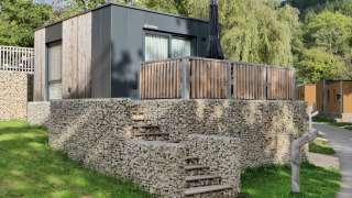 Modern tiny house with wooden exterior, stone steps, and patio, surrounded by greenery and lush trees.
