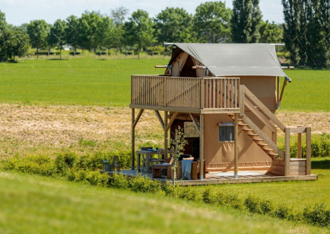 Une tente de glamping moderne avec balcon supérieur et coin salon extérieur, dans un paysage rural verdoyant.