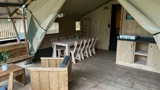 Binnenkant van een safaritent met houten meubilair, eettafel, zithoek en kleine kitchenette.
