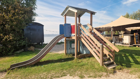 Speeltuin met glijbaan en houten hut bij een glamping, met tent en water op de achtergrond.