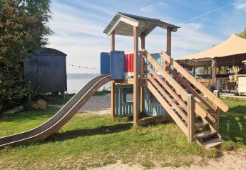 Parco giochi con scivolo e casetta di legno presso una glamping, con tenda e acqua sullo sfondo.