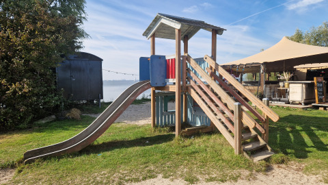 Spielplatz mit Rutsche und Holzhütte bei einer Glamping-Unterkunft, Zelt und Wasser im Hintergrund.