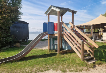Speeltuin met glijbaan en houten hut bij glamping, met tent en water op de achtergrond.